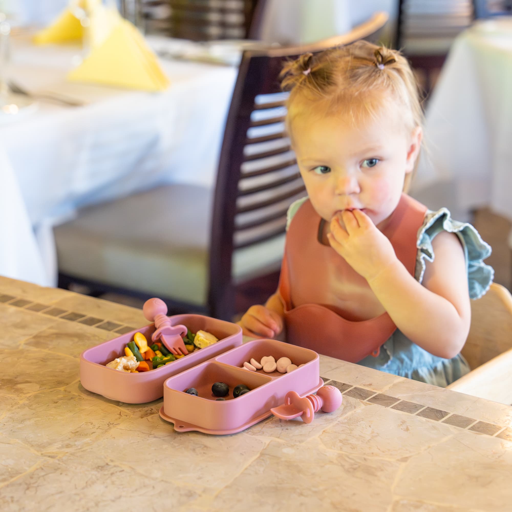 Toddler girl in pink bib eats from a pink silicone divided container at a table.