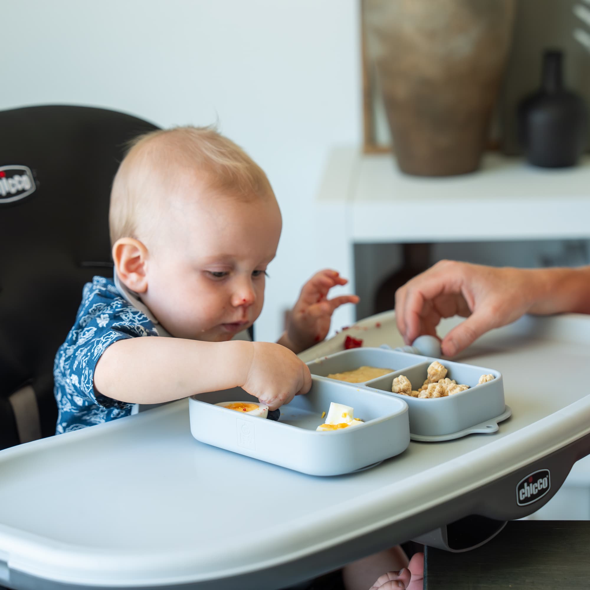 A baby in a high chair eats finger foods from a grey divided silicone tray.