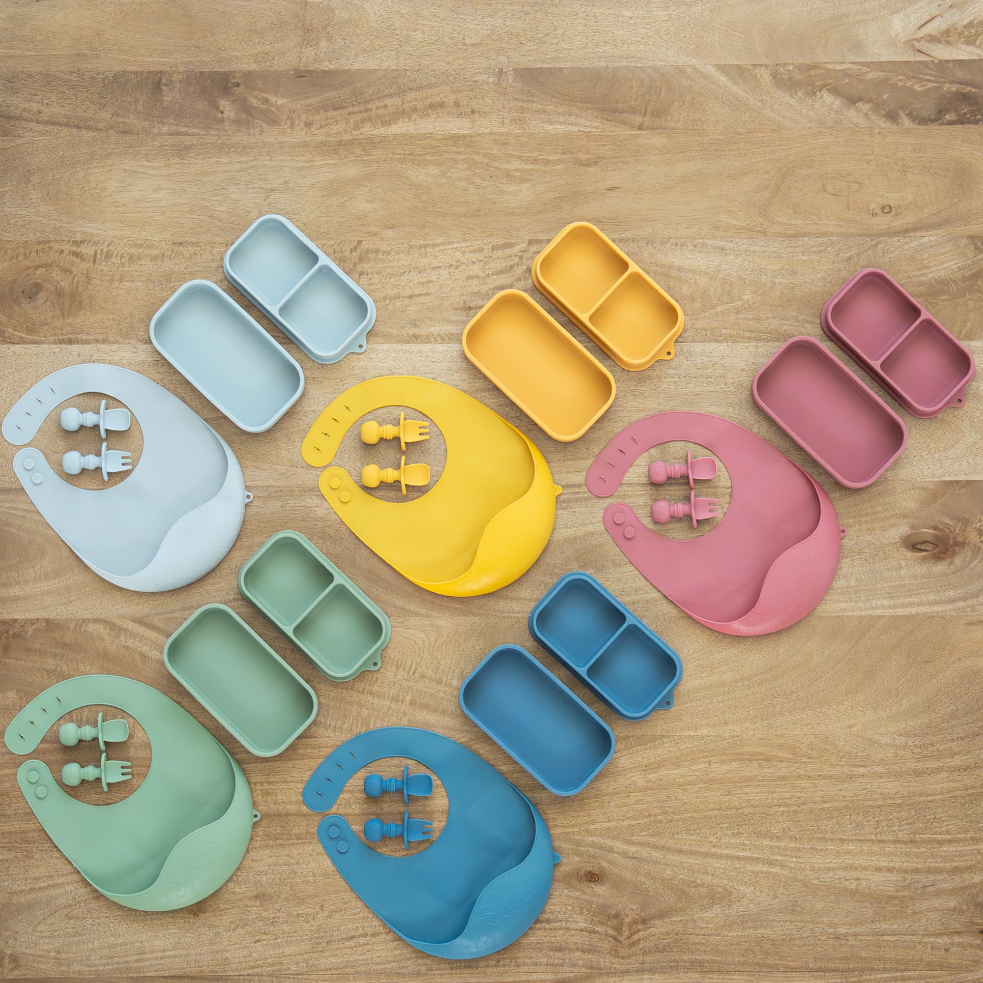Five colorful silicone baby feeding sets with bibs, utensils, and snack containers on wood.